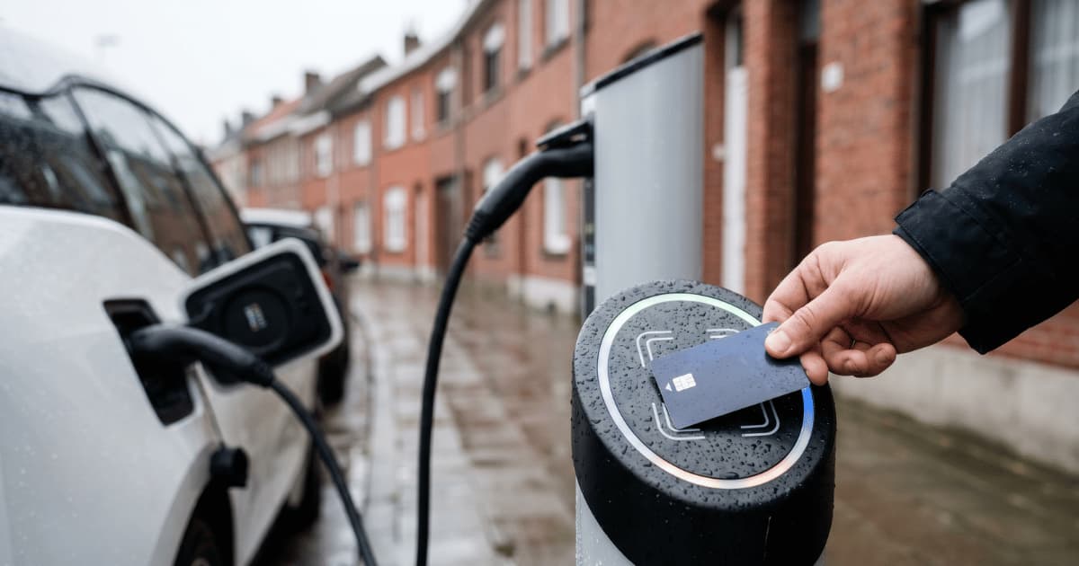 Une main scanne une carte de recharge sur le lecteur RFID d'une borne publique pendant qu'une voiture électrique blanche se recharge dans une rue résidentielle belge en briques sous la pluie.
