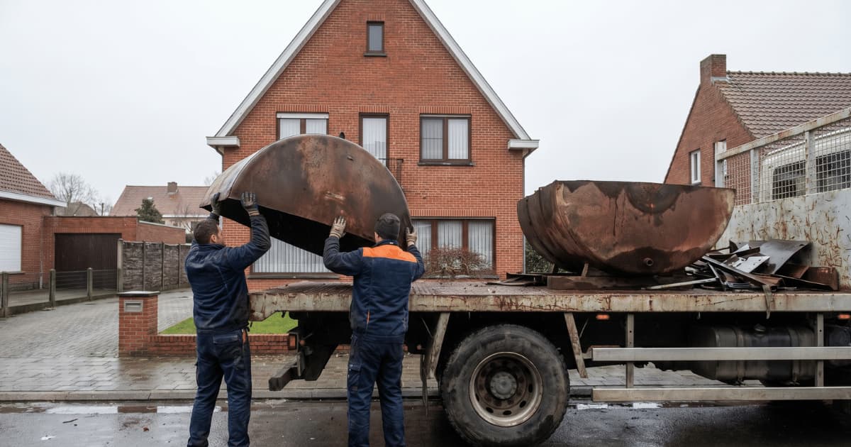 Deux ouvriers chargent les sections découpées d'une ancienne citerne à mazout sur le plateau d'un camion stationné devant une habitation belge en brique rouge pour évacuation vers un centre de traitement agréé.