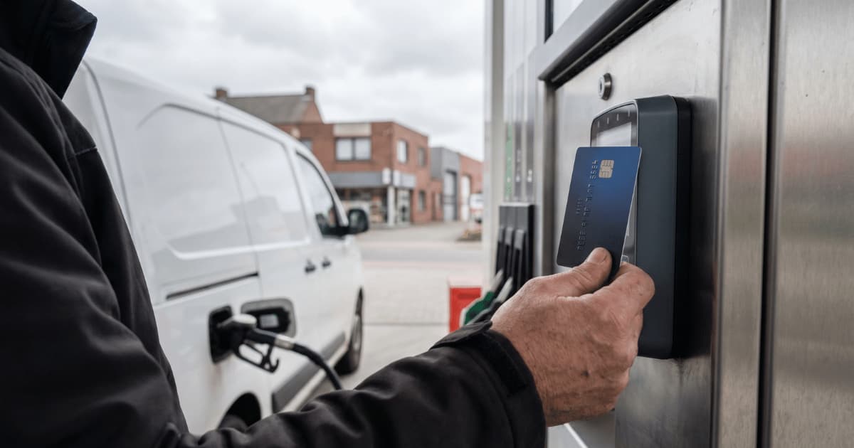 Un chauffeur en veste noire présente sa carte carburant bleue contre le terminal de paiement d'une pompe à essence tandis que sa camionnette blanche est en cours de ravitaillement sur un forecourt belge sous un ciel couvert.