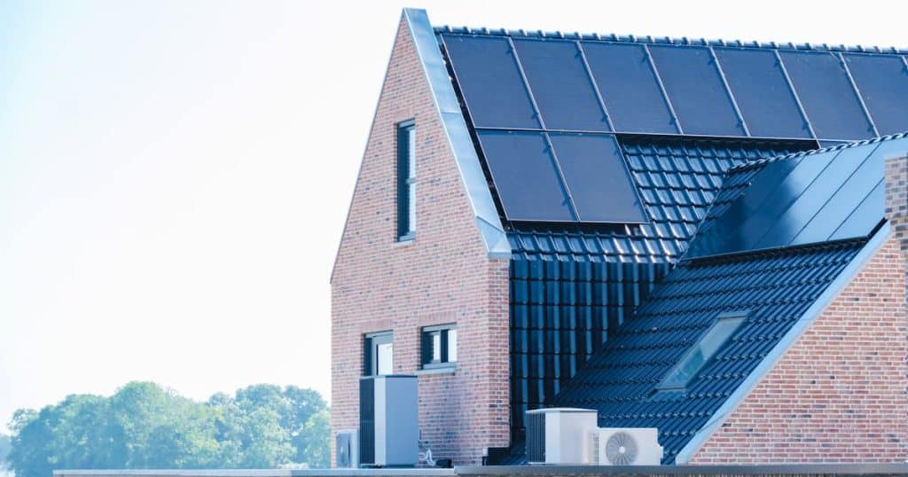 Une maison avec une façade grise, l'unité extérieure d’un système de climatisation et un toit gris équipé de panneaux solaires.