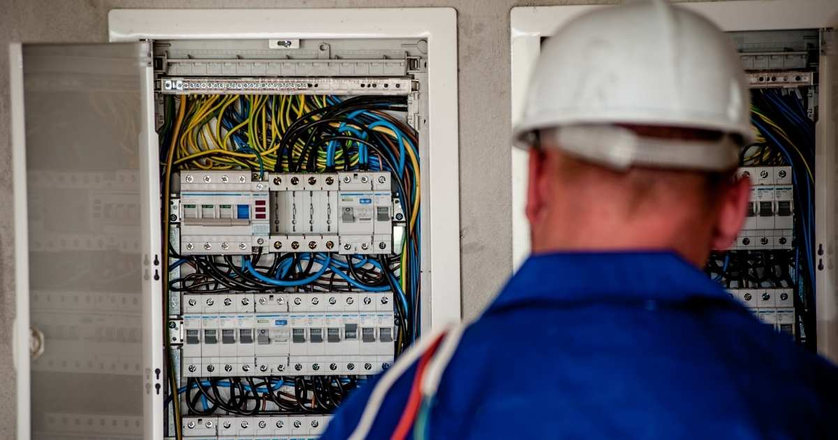 Un contrôleur agréé en combinaison bleue et casque de sécurité blanc inspecte les tableaux électriques d’un appartement.