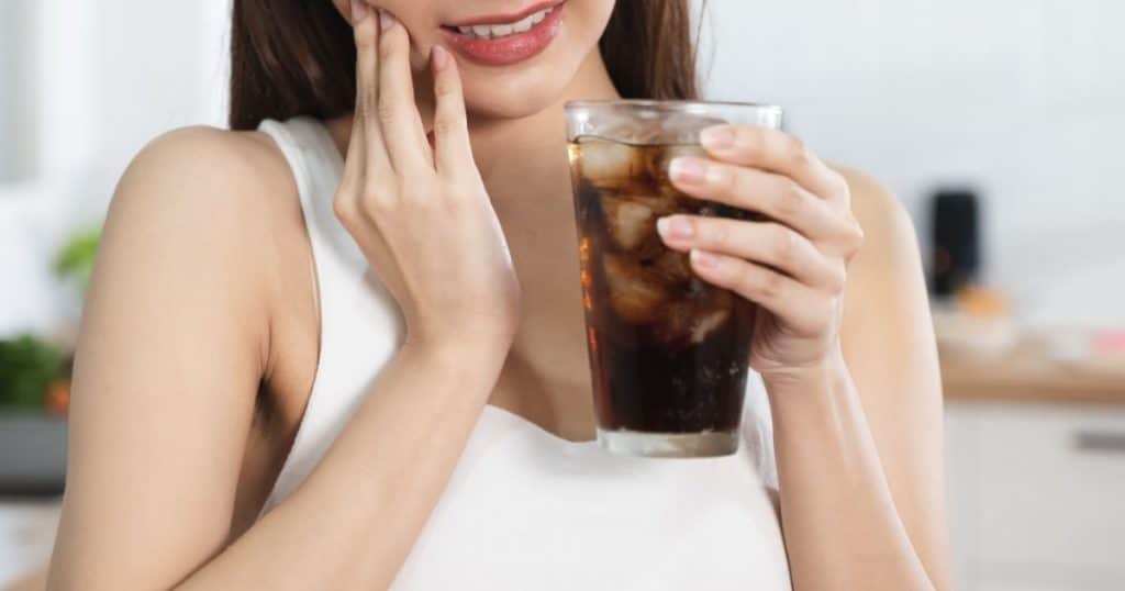 Une jeune femme tient un verre de soda avec des glaçons dans une main et se tient la mâchoire de l'autre. 