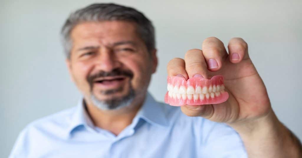 Un homme souriant tient un dentier entre ses mains. 