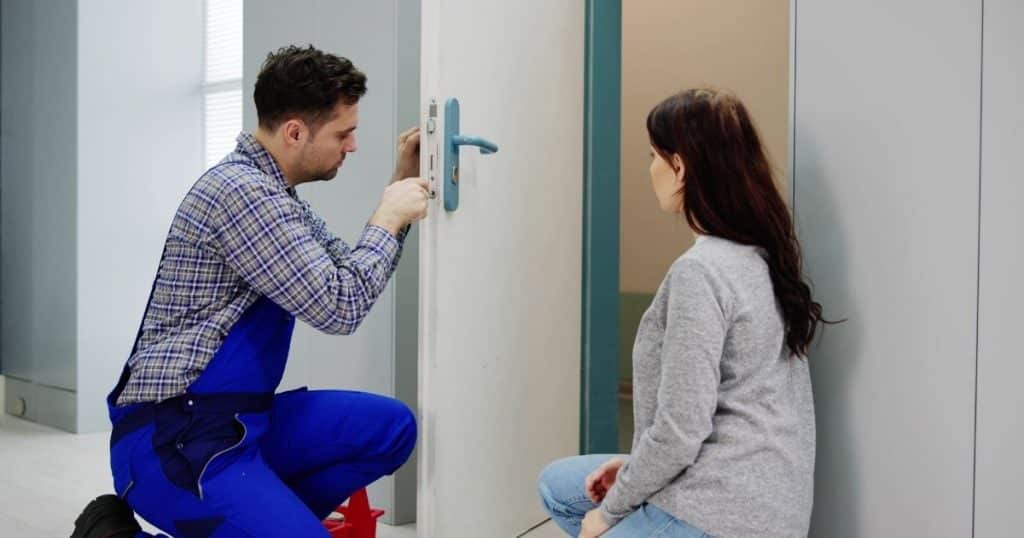 serrurier en bleu de travail en train de réparer le cylindre d'une porte avec une cliente qui le regarde