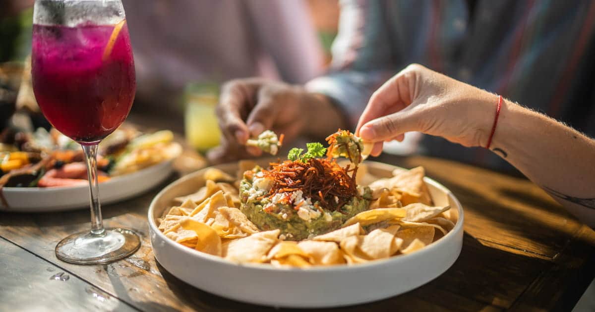 Deux personnes partagent un plat de guacamole garni de légumes et accompagné de chips de maïs, posé sur une table en bois, avec un cocktail violet en premier plan.