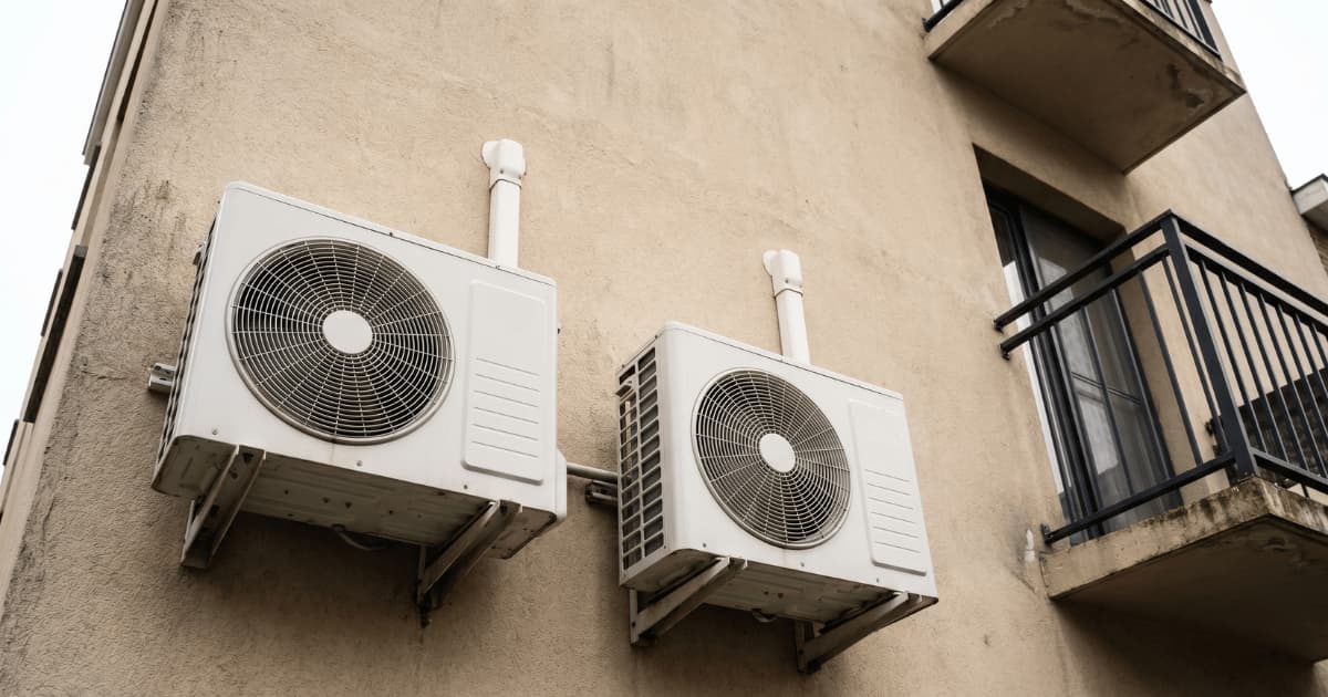 Deux unités extérieures de climatisation blanches fixées sur des supports métalliques contre la façade beige d'un immeuble belge, vues en contre-plongée avec des conduites blanches qui remontent vers le mur et un balcon visible à droite.
