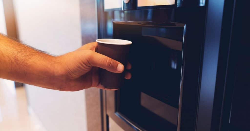 Distributeur automatique de café avec un homme en train de tendre son gobelet en carton pour se servir un café au bureau