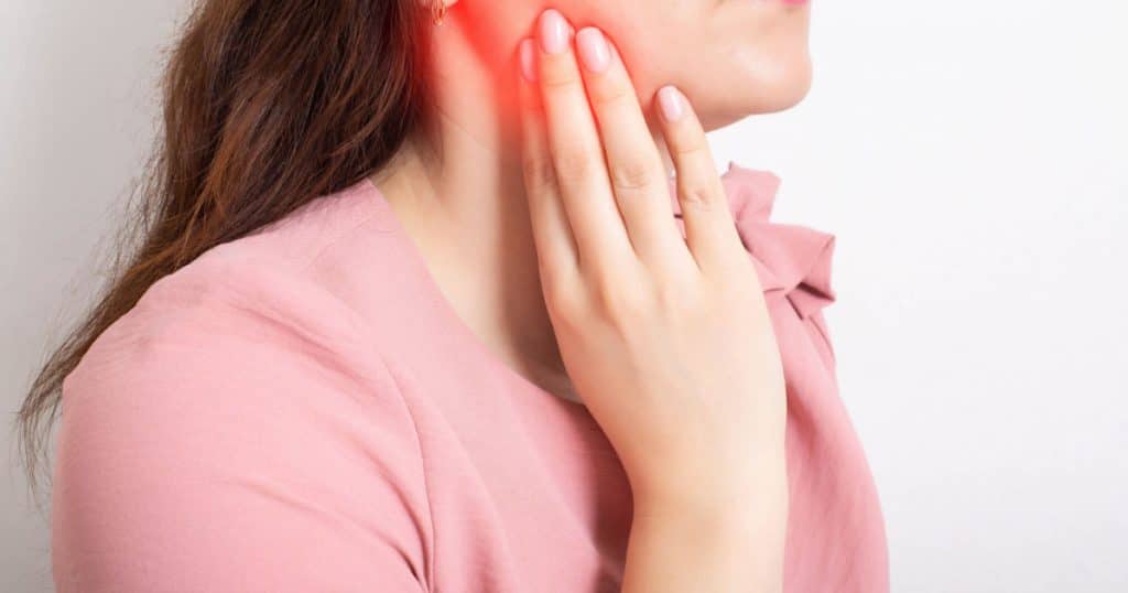Une jeune femme tient sa mâchoire douloureuse et artificiellement rouge avec sa main. 