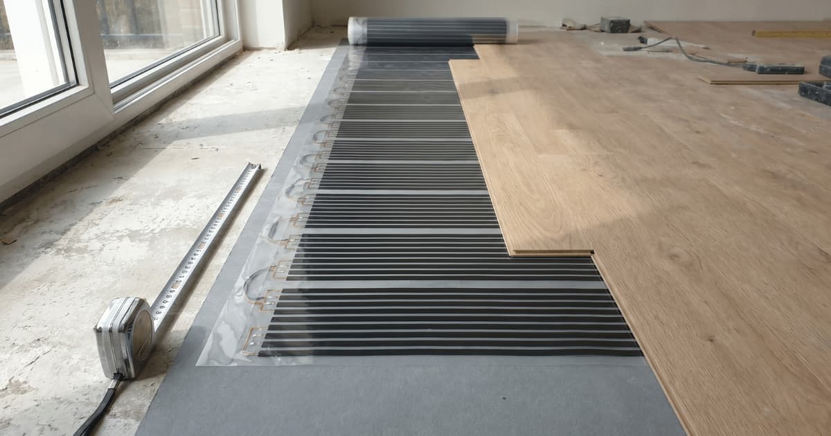 Film chauffant infrarouge à bandes de carbone déroulé sur une chape brute avec des lames de parquet clair partiellement posées par-dessus et un mètre ruban au sol.