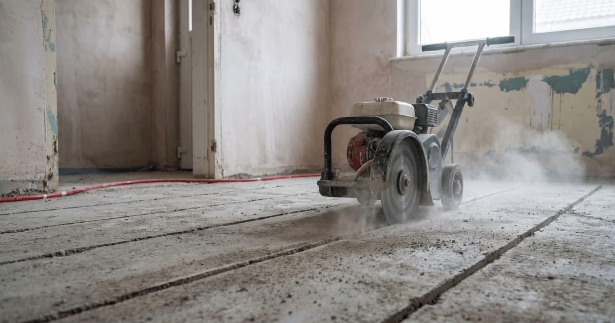 Fraiseuse thermique en action creusant des rainures parallèles dans une chape existante pour y loger des tuyaux de chauffage au sol dans une maison en rénovation avec de la poussière de coupe visible.