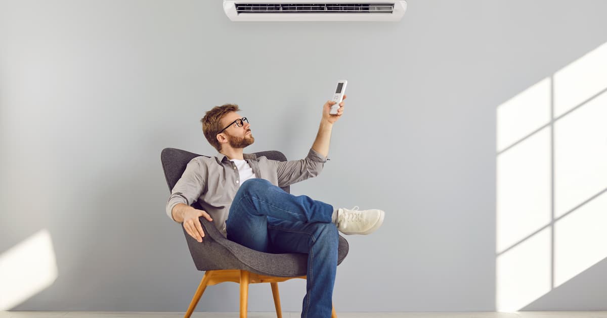 Jeune homme barbu en lunettes et vêtements décontractés allumant l'air conditionné assis sur sa chaise dans un espace vide jouissant d'air frais et climatisé à l'aide d'une télécommande isolée sur fond gris du mur.