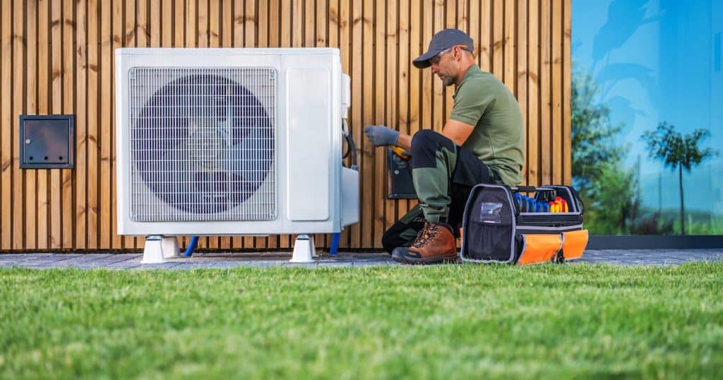 Un technicien assure l'entretien d'un climatiseur situé à côté d'une maison moderne. Habillé en tenue de travail, il utilise les outils d'une boîte à outils organisée tout en étant assis sur l'herbe. 