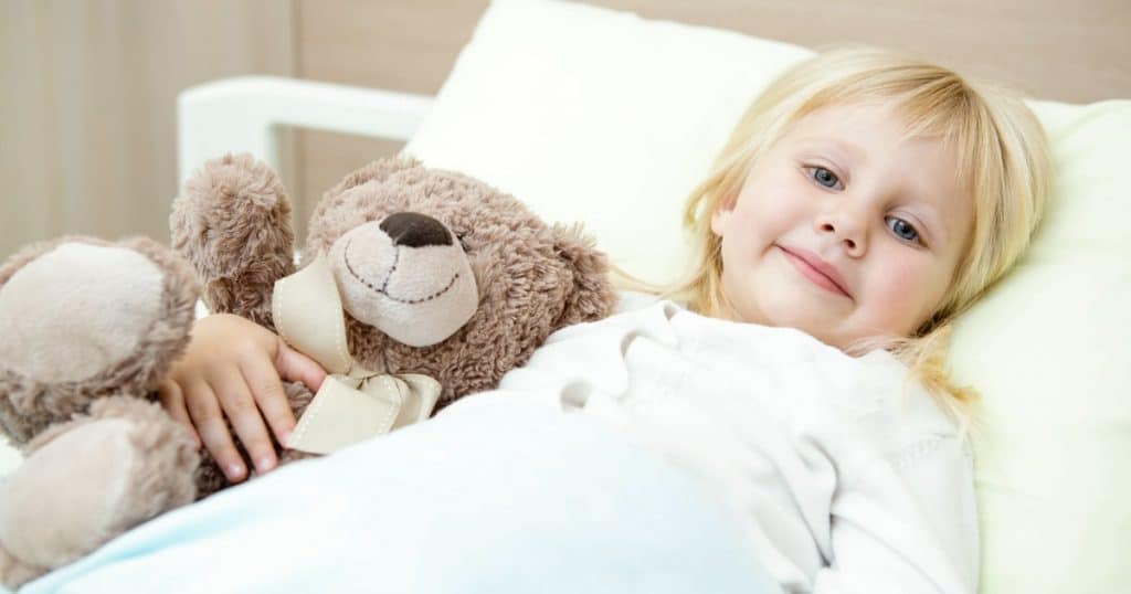 Une enfant souriante sur son lit d'hôpital tient un ours en peluche entre les bras.
