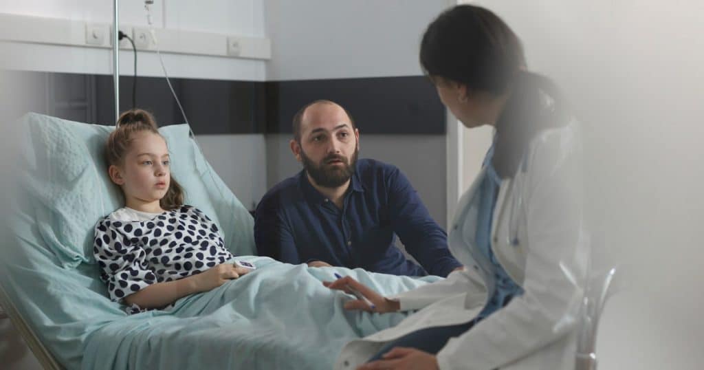 Une enfant adossée sur son lit d'hôpital avec son père à ses côtés parlent à une docteur en blouse blanche. 