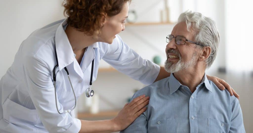 Une femme en blouse blanche avec un stéthoscope autour du cou rassure un patient souriant, les mains sur ses épaules. 