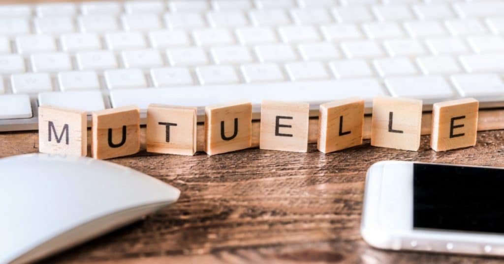Sur un bureau, aux côtés d'un clavier, d'une souris et d'un smartphone sont posées des lettres en bois de Scrabble qui forment le mot "mutuelle". 