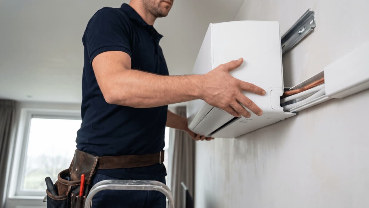 Gros plan en contre-plongée d'un installateur qui clipse une unité intérieure de climatisation blanche sur sa plaque de montage métallique, les raccords en cuivre visibles derrière l'appareil.