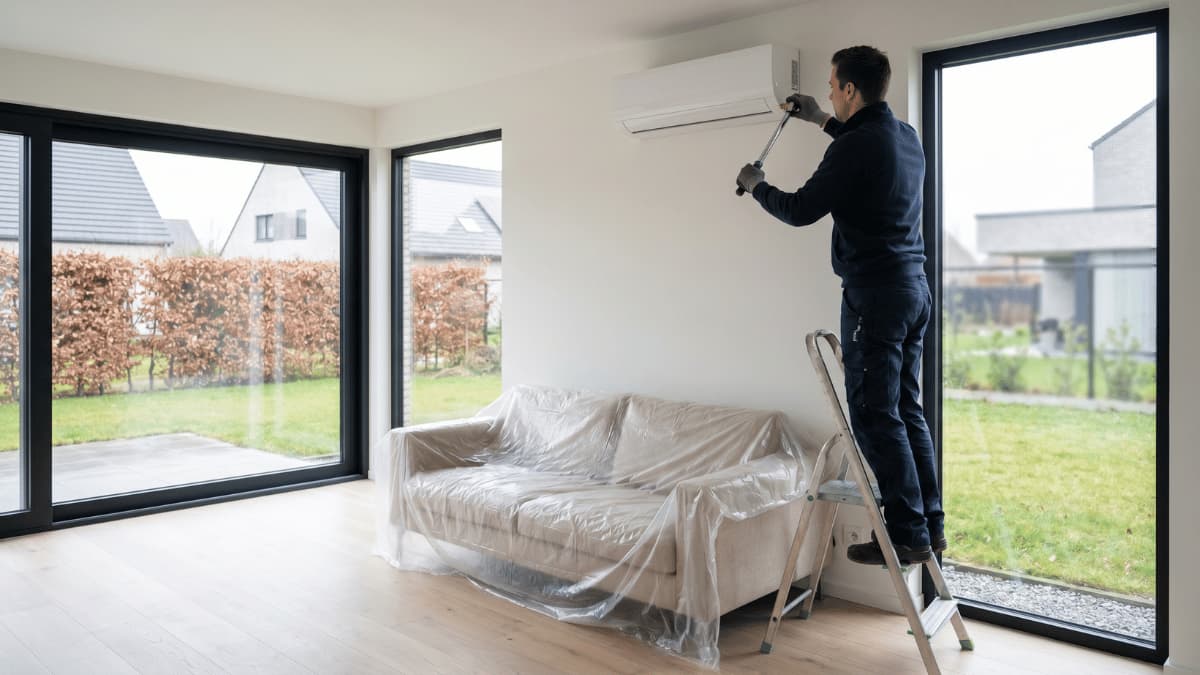 Un installateur en combinaison bleu foncé se tient sur un escabeau dans un salon lumineux aux grandes baies vitrées et place une unité intérieure de climatisation murale blanche sur le mur, tandis que le canapé est protégé par une bâche plastique.