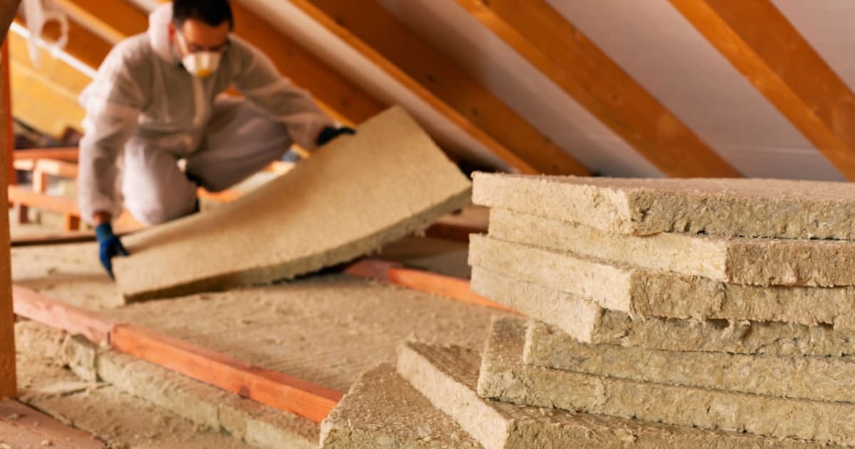 Un homme en combinaison de protection isole les combles d'une maison avec des plaques d'isolant.