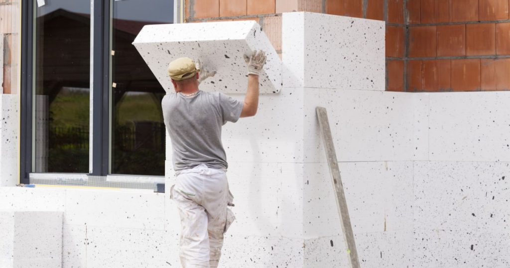 Un artisan installe un bloc d'isolant sur la façade extérieure d'un bâtiment. 