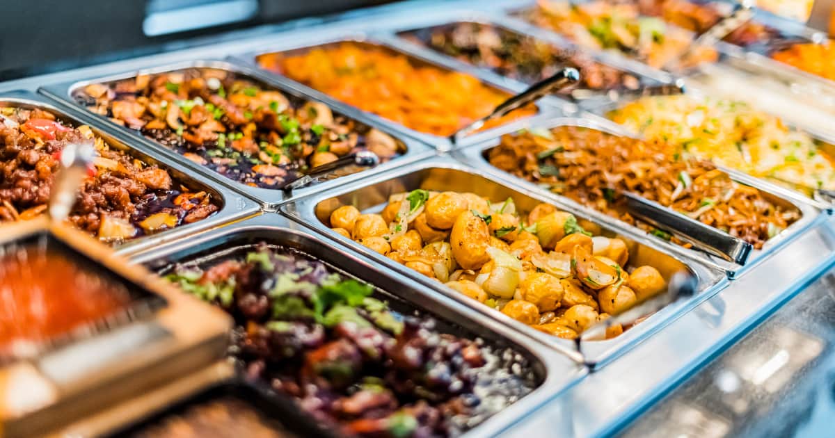 Buffet de plats chauds variés, comprenant pommes de terre rôties, nouilles sautées, légumes et viandes en sauce, présentés dans des bacs en inox.