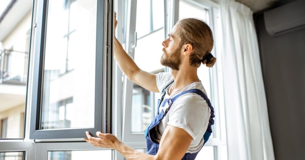 Homme installant une fenêtre moderne en aluminium dans un appartement lumineux, portant une salopette de travail et un t-shirt blanc
