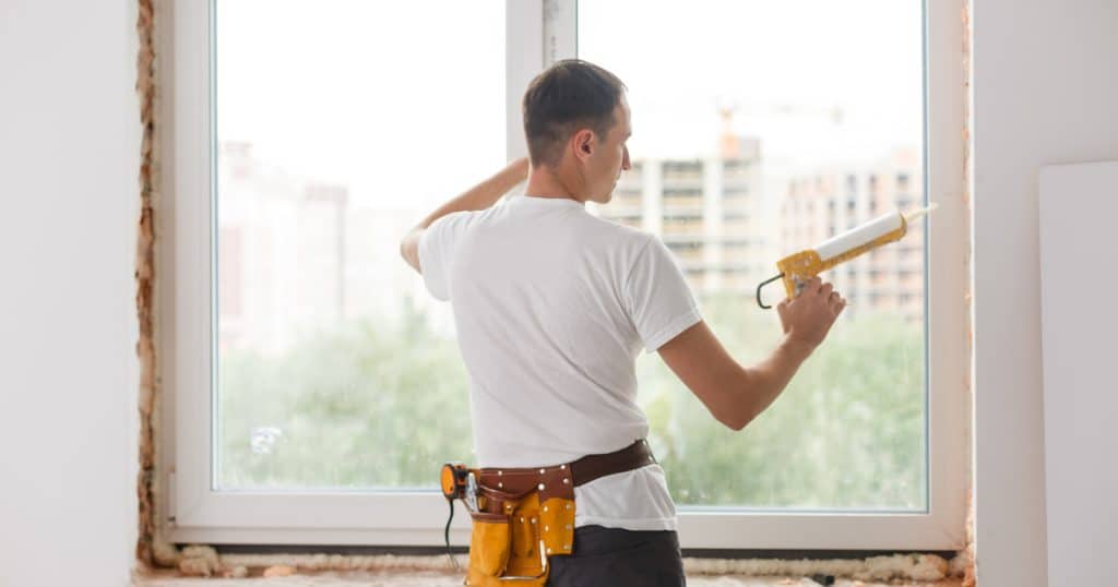 Installateur appliquant du mastic autour d’un châssis de fenêtre en PVC, équipé d’une ceinture à outils, dans un appartement en cours de rénovation