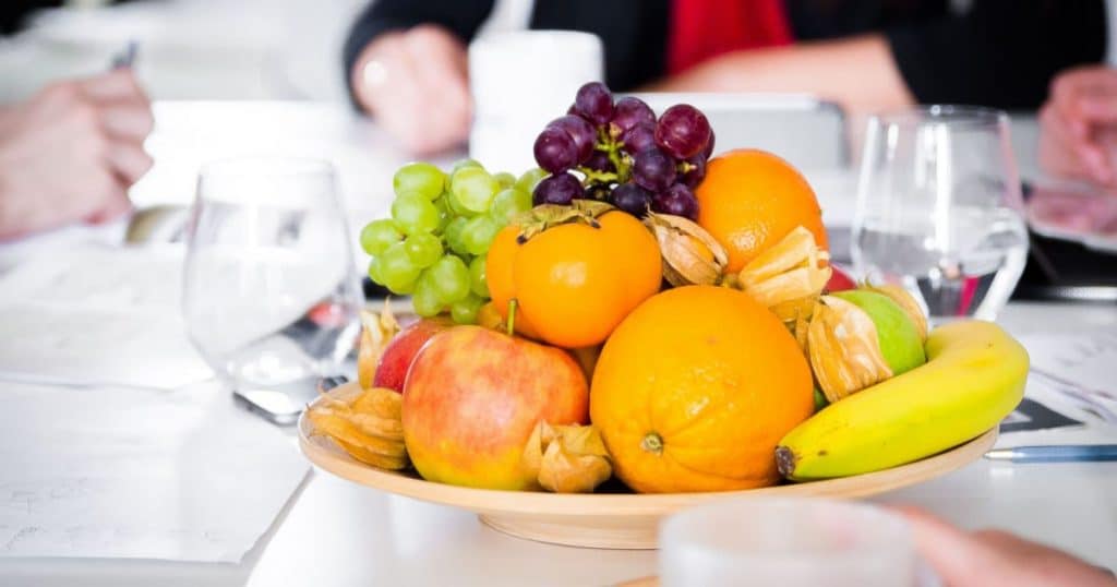 corbeille de fruits variés contenant des pommes, oranges, kaki, banane,... posée sur la table d'une salle de réunion avec différents collègues autour en train de travailler
