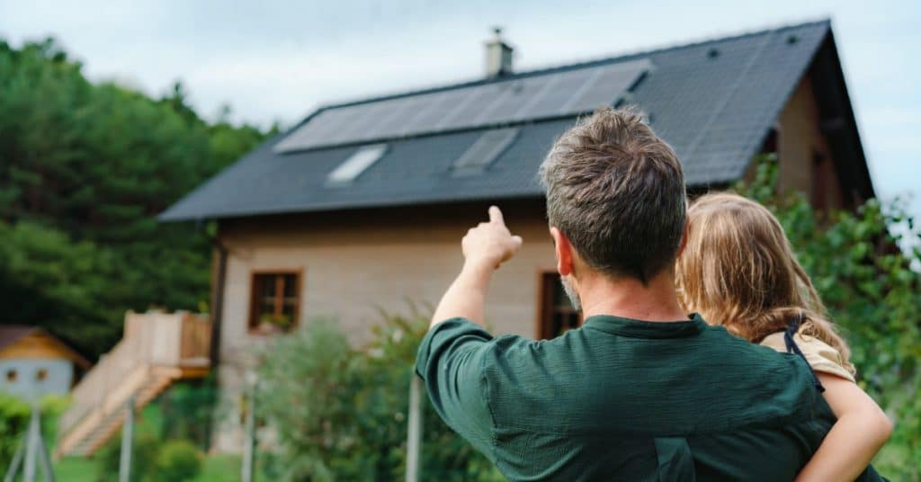 un homme tient sa fille dans ses bras et pointe vers le toit de la maison où se trouvent des panneaux solaires