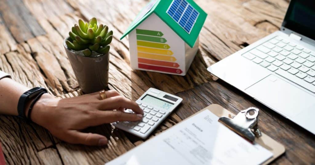 Une table montre un document, un ordinateur portable, une main qui tape sur une calculatrice et un modèle réduit de maison avec les jauges PEB d'évaluation des performances énergétiques. 