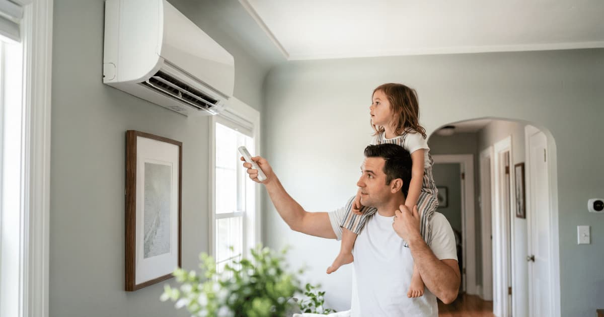 Un père en t-shirt blanc porte sa fille sur les épaules et pointe une télécommande vers une unité de climatisation murale blanche dans un salon aux murs vert-gris, avec un cadre décoratif et une plante verte au premier plan.