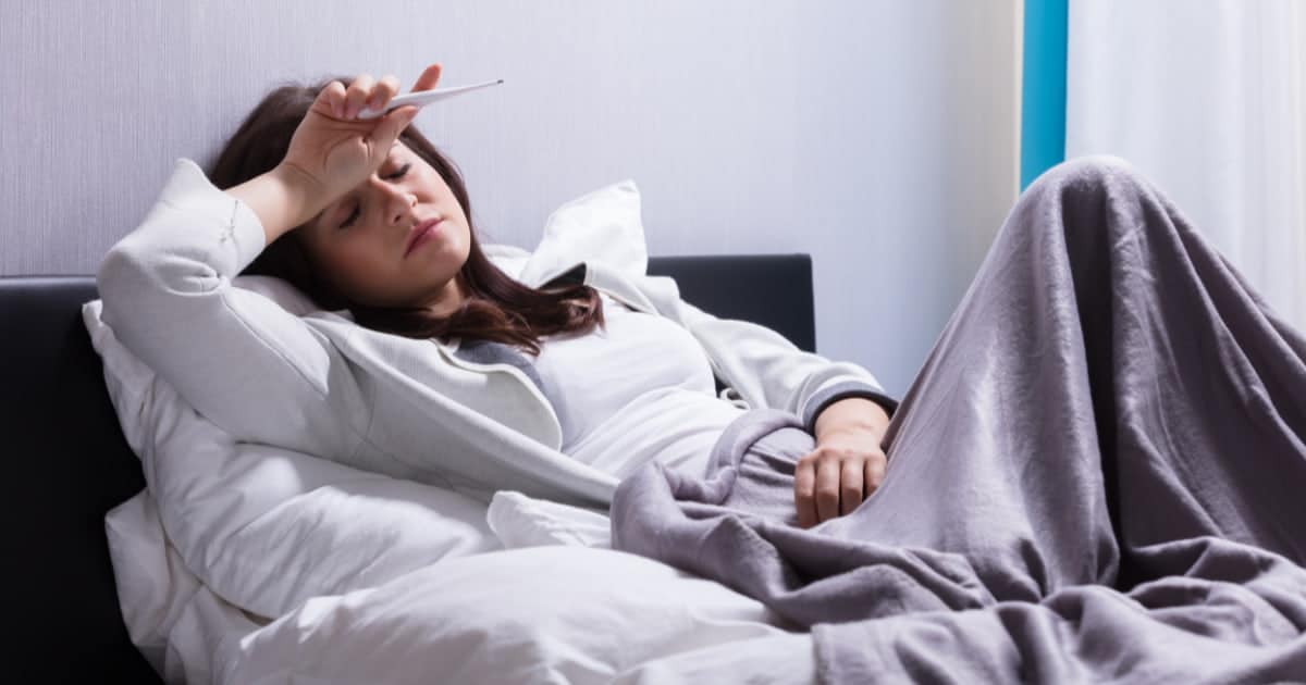 Femme allongée dans un lit, l’air malade, tenant un thermomètre sur son front, couverte d’une couverture grise.