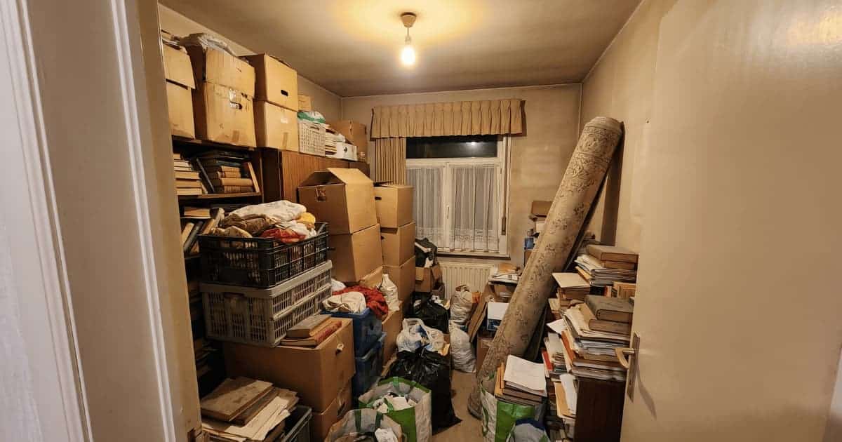 Une petite pièce déborde de cartons empilés jusqu'au plafond, de caisses en plastique, de sacs et de piles de livres tandis qu'un tapis roulé s'appuie contre le mur sous une ampoule nue.