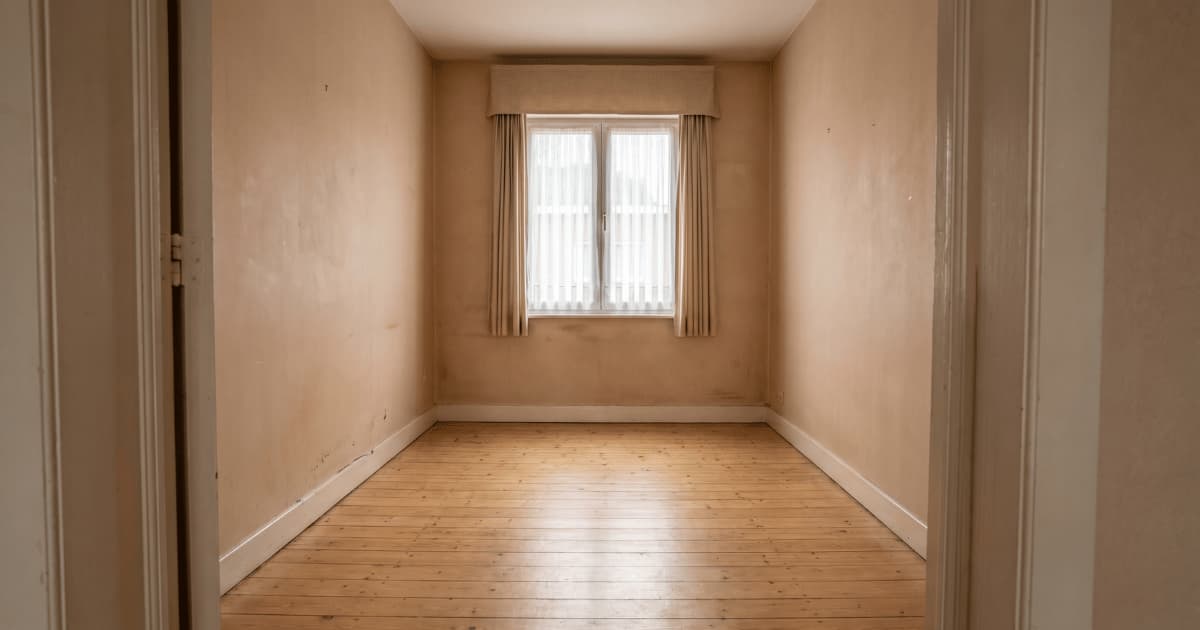 Une petite pièce entièrement vidée révèle un parquet en bois clair et des murs beiges défraîchis avec une fenêtre habillée de rideaux et un radiateur au fond.