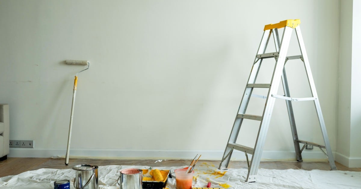 Salle préparée pour la peinture avec une échelle, des boîtes de peinture, rouleau, et des brosses sur un tissu goutte à goutte. Les murs sont nus, prêts pour une nouvelle couche de peinture.