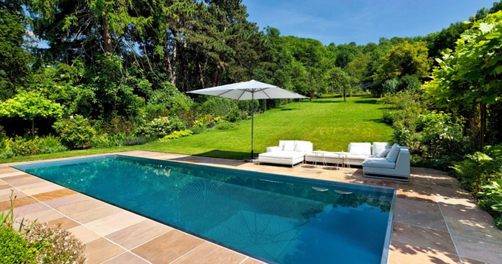 Piscine creusée dans un jardin avec terrasse en carrelage beige, meubles extérieurs et parasol.