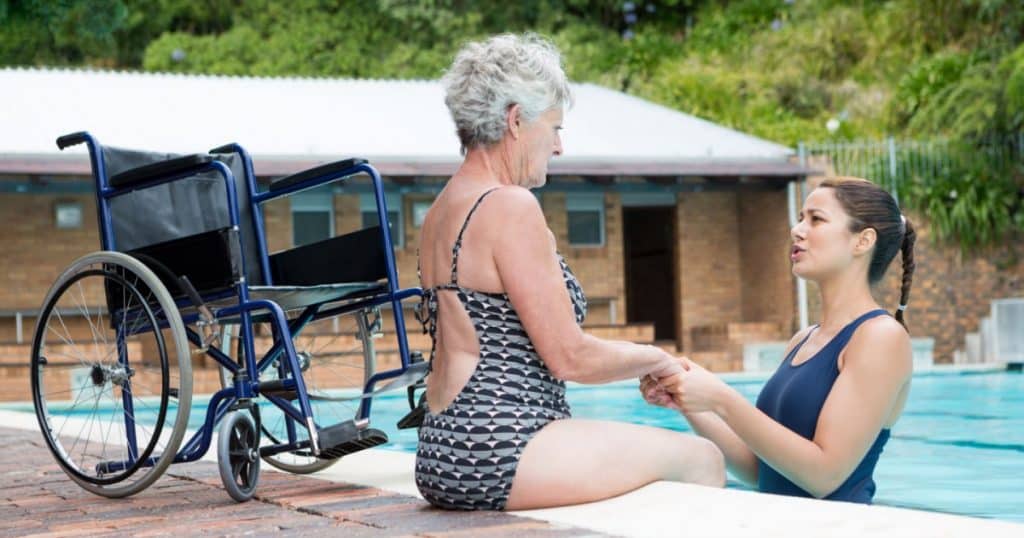 Dame handicapée assise sur le bord de la piscine, tenant les mains d'une jeune femme brune qui est dans la piscine. 