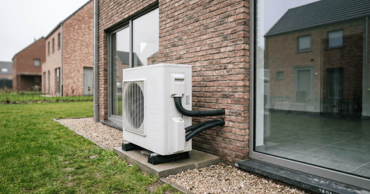 Unité extérieure blanche d'une pompe à chaleur air-eau posée sur un socle en béton avec bande de gravier le long d'une façade en brique brune d'une maison moderne dans un quartier résidentiel belge par temps couvert.