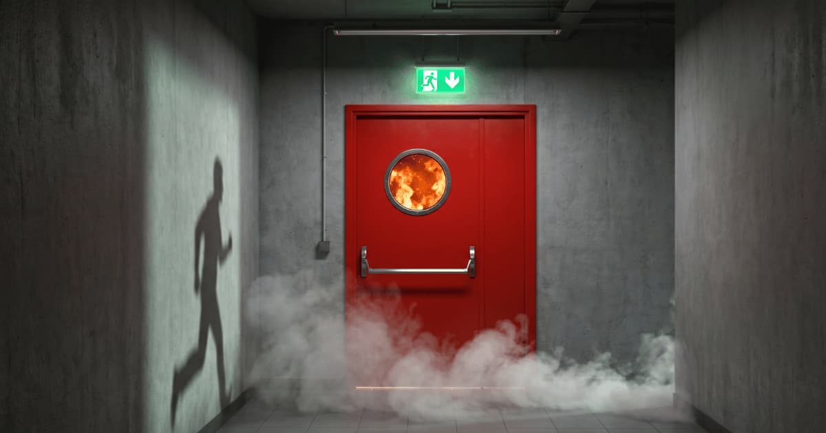 Une porte coupe feu rouge munie d'un hublot rond laisse entrevoir des flammes vives de l'autre côté tandis que de la fumée blanche se répand au sol d'un couloir en béton brut où un panneau de sortie de secours vert brille au dessus de la porte et l'ombre d'une personne en fuite se projette sur le mur gauche.