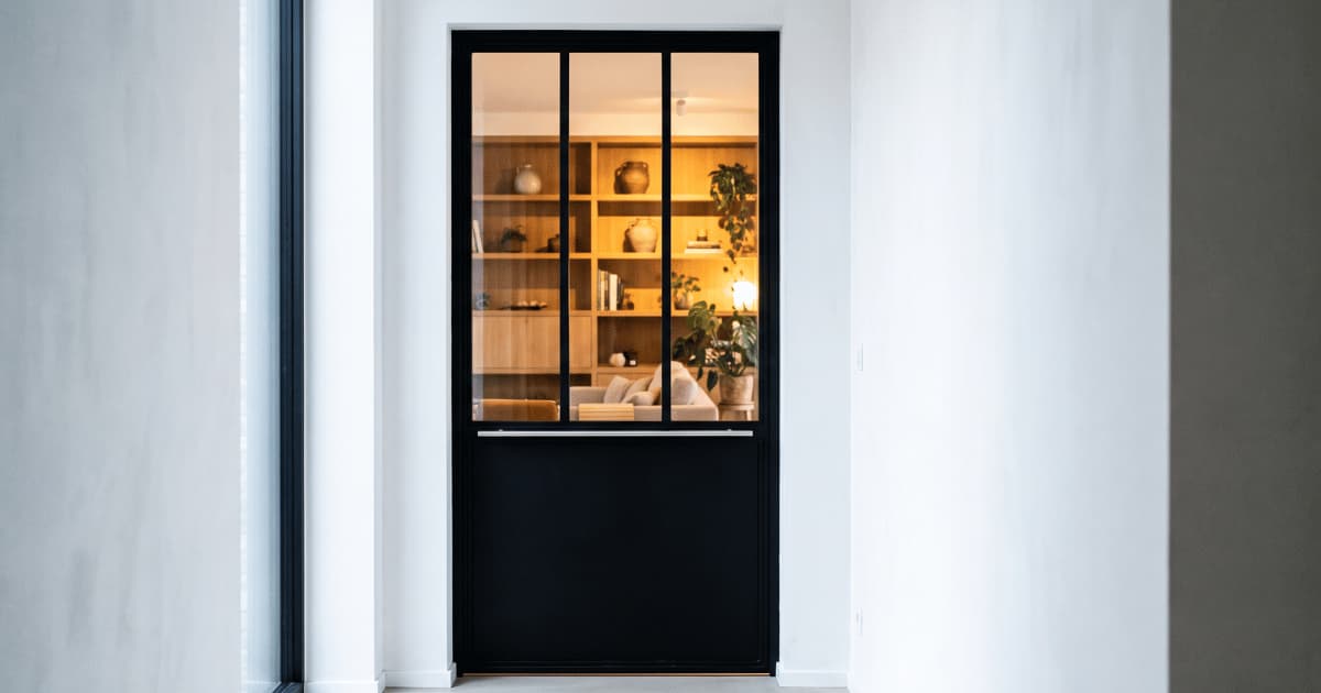 Une porte intérieure de style atelier avec un cadre en aluminium noir mat et trois panneaux vitrés verticaux séparés par des montants fins laisse entrevoir un salon chaleureusement éclairé avec une grande bibliothèque en bois garnie de vases et de plantes vertes tandis qu'une barre de poignée horizontale en métal traverse la porte au dessus du panneau inférieur plein noir le tout encastré dans un mur blanc lisse baigné de lumière naturelle.