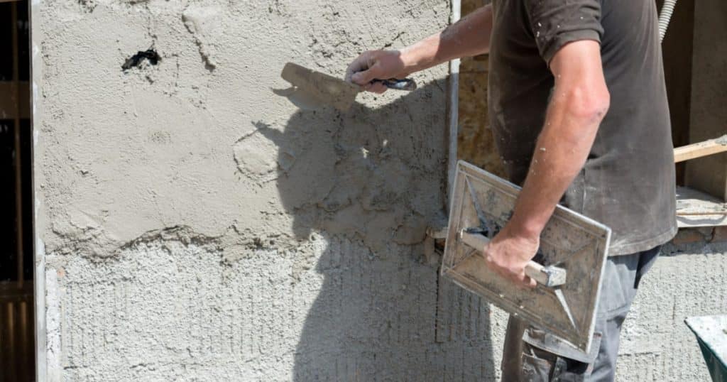 Un homme muni d'une taloche et d'une truelle pose un enduit extérieur de type crépi sur la façade d'une maison. 