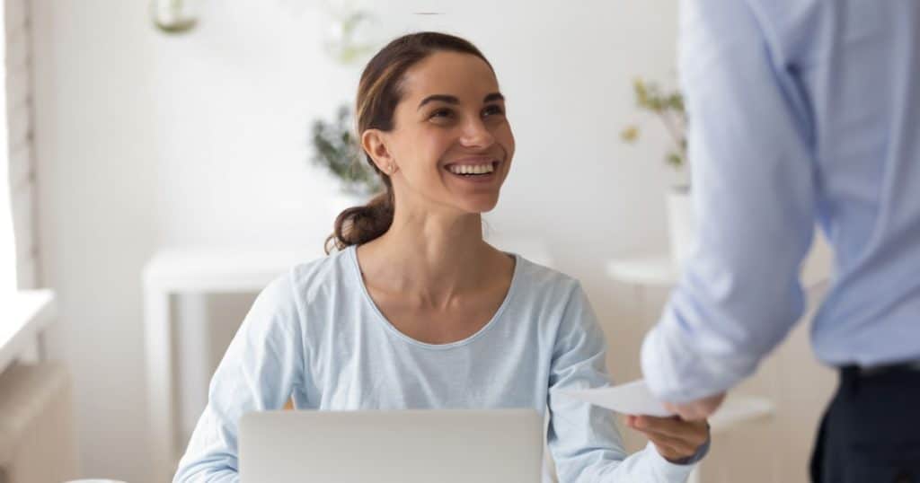 Une femme assise à son bureau devant un ordinateur portable sourit à un homme qui lui donne une enveloppe. 