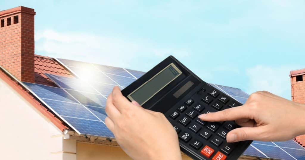 Des mains qui utilisent une calculatrice se détachent sur le toit d'une maison recouvert de panneaux photovoltaïques.