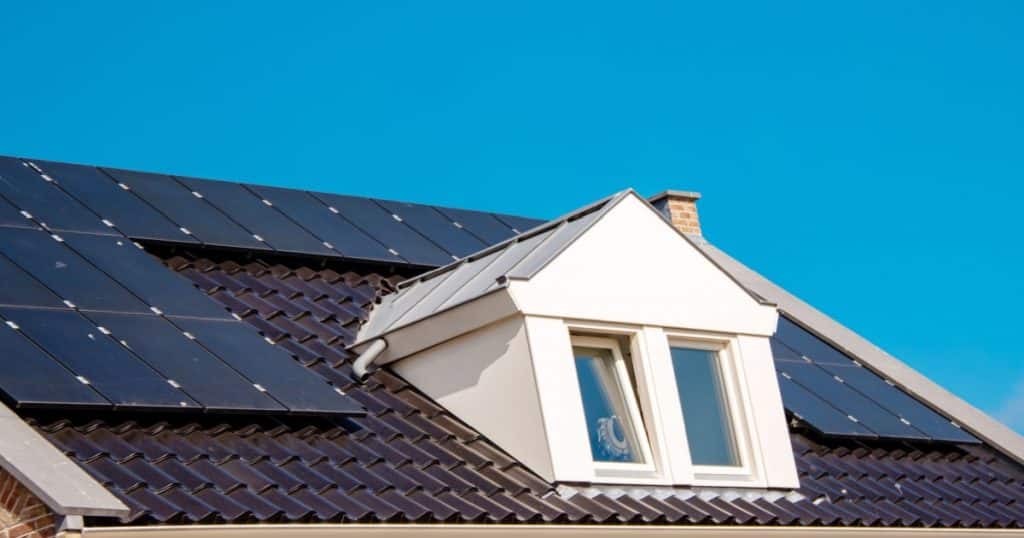 Le toit d'une maison avec une fenêtre recouvert de panneaux photovoltaïques se détache sur un ciel très bleu. 
