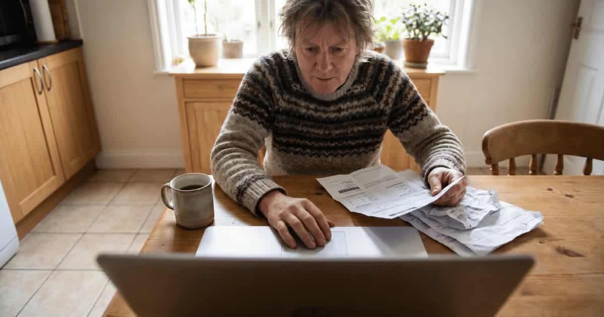 Un propriétaire en pull tricoté consulte des factures d'énergie à sa table de cuisine en bois tout en posant une main sur le trackpad de son laptop ouvert, une tasse de café à côté de lui et des documents froissés étalés sur la table.