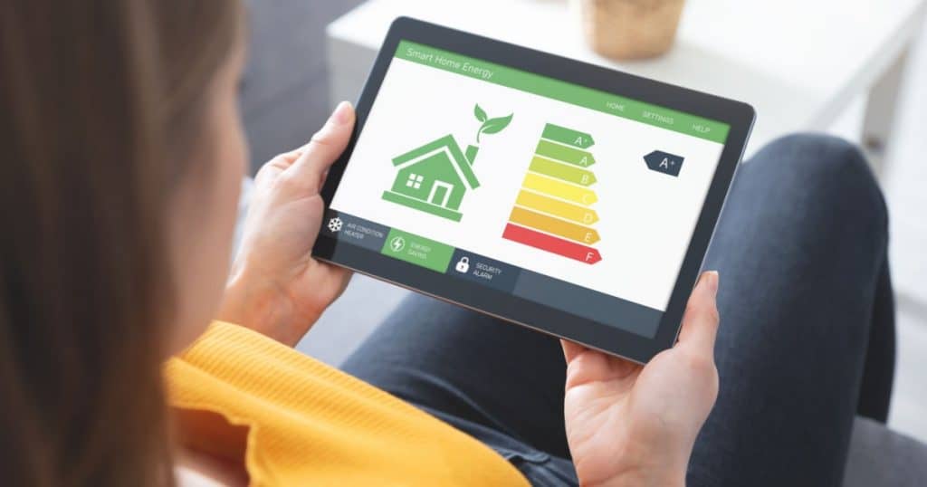 Une femme assise dans son salon tient dans ses mains une tablette tactile qui affiche le label énergétique de son logement.