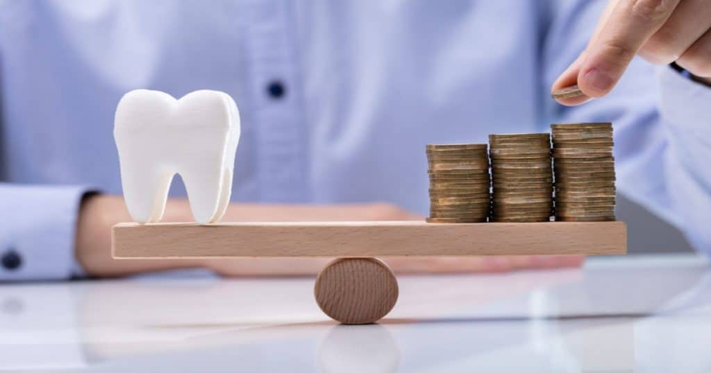 Une main ajoute une pièce sur un tas d'autres, sur une petite planche en bois en équilibre, une grosse dent en plastique se trouve de l'autre côté de la planche.