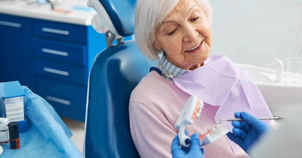 Une personne âgée est à un rendez-vous chez le dentiste et ce dernier lui montre une maquette des arcades inférieure et supérieure. 