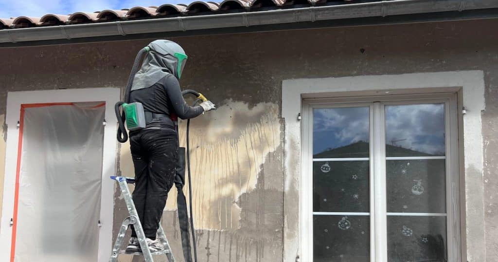 Personne en tenue protectrice décapant une façade en béton via une sableuse