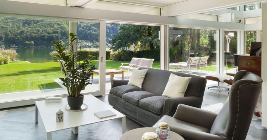 salon avec canapés gris installés dans un véranda donnant sur un jardin avec une pelouse et un lac.